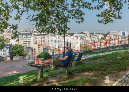 Un homme est assis sur un banc de parc avec ses pieds en regardant son téléphone cellulaire avec une vue sur la ville en arrière-plan, à Lisbonne, au Portugal. Banque D'Images