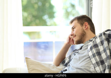 Portrait of a smiling man sitting on a couch et à l'extérieur à travers une fenêtre dans la salle de séjour à la maison Banque D'Images