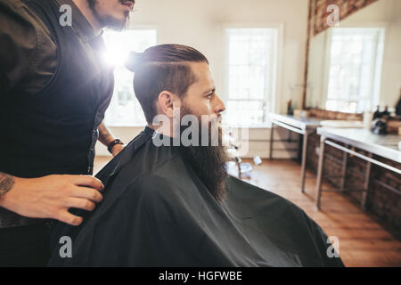 Vue de côté tourné de beau barbu dans une cape noire la coupe de cheveux dans le barbershop, avec coiffure par permanent. L'homme assis à un salon de coiffure.