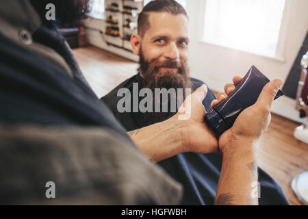 Salon de coiffure au service de son client. L'homme à barbe sitting in chair et regardant hairstylist. L'accent sur coiffure holding crème cosmétique dans la main. Banque D'Images