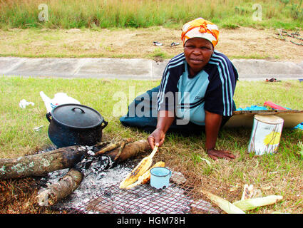 Maïs bouilli ou grillé à vendre dans une opération de fortune sur route, Afrique du Sud. Une femme locale assise sur le sol, une vue commune partout. Banque D'Images