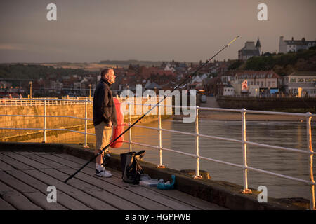 Éclairé par le soleil du soir au coucher du soleil (mais sous un ciel sombre) est un homme, pêche en mer à partir de la jetée Ouest - Whitby, North Yorkshire. Banque D'Images