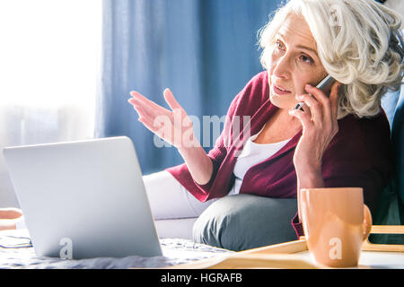 Attractive senior Woman talking on smartphone pendant que lying on bed Banque D'Images