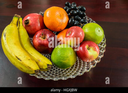 Fruits mélangés. banana raisins noirs grenade rouge orange lime douce Apple dans un bol en verre avec fond de bois. Banque D'Images