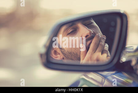 Miroir côté réflexion sur le bâillement fatigué fatigué endormi épuisé jeune homme au volant de sa voiture dans la circulation après de longues heures de route. Banque D'Images