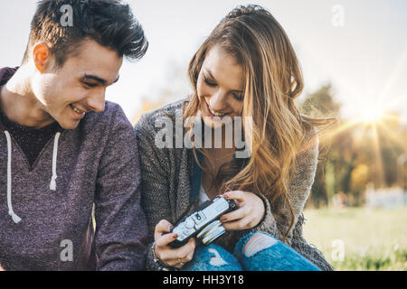 Happy smiling young couple outdoors assis sur l'herbe et la tenue d'un appareil photo numérique, sur fond de bois Banque D'Images