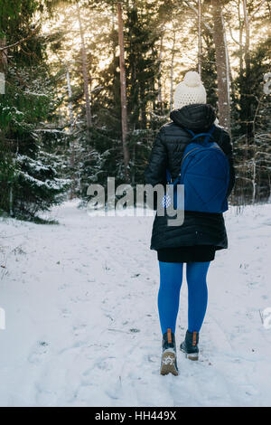 Young woman walking in winter forest, coucher de la lumière à travers les arbres enneigés. Vue arrière Banque D'Images