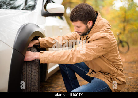 Jeune homme barbu contrôle pneu de voiture en forêt d'automne Banque D'Images