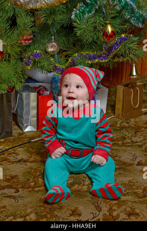 Smiling little baby boy en costume vert-rouge est assis sur le plancher tapis napless couvrant près de l'arbre de Noël. Banque D'Images