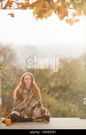 Serene woman wrapped in blanket méditant sur patio d'automne Banque D'Images