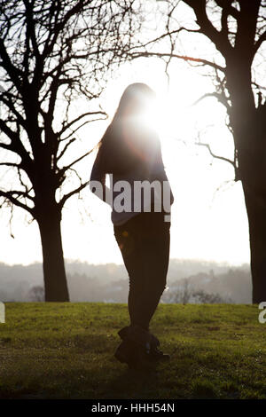 Silhouette d'une vue arrière d'un seul femme méconnaissable par un banc dans un endroit calme avec un soleil ciel et les arbres. Banque D'Images