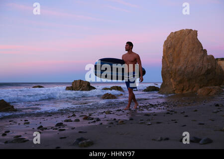 Hispanic man carrying surfboard on beach at sunset Banque D'Images