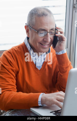 Smiling Hispanic man multi-tâches avec téléphone cellulaire et ordinateur portable Banque D'Images
