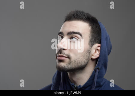 Pensive young bearded man looking up wearing hooded shirt bleu sur fond gris. Banque D'Images