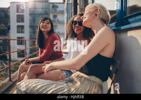 Trois jeunes amis féminins assis sur un balcon et s'amusant. Les femmes de détente en plein air et le chat. Banque D'Images