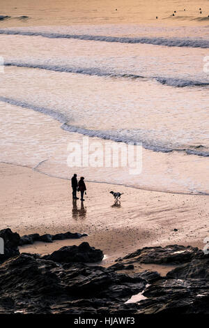 Deux personnes et leur chien vu en silhouette alors que le soleil se couche sur la plage de Fistral, Newquay, Cornwall. Banque D'Images