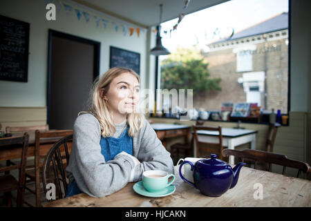 Jeune femme assise dans un café, une tasse de thé et une théière sur la table, l'air pensif Banque D'Images