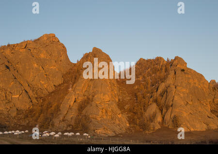 Un camp avec plusieurs yourtes blanches à la base des montagnes de pierre robuste qui sont d'or au coucher du soleil. Situé en Mongolie. Banque D'Images