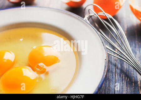 Trois oeufs crus dans une plaque de métal blanc sur une table, avec quelques coquilles d'œufs fêlés et batteur à oeufs Banque D'Images