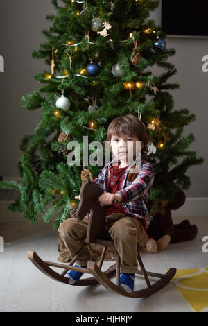 Little Boy riding un jouet cheval de bois à côté d'un arbre de Noël et de rires Banque D'Images