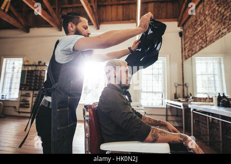 Mettre le cap coiffure coiffeur au client avant de couper les cheveux. Coiffure masculine au travail avec bel homme assis sur une chaise. Banque D'Images