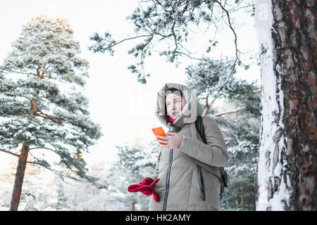 Cheerful woman texting on smartphone orange lors d'un voyage à la forêt en hiver. Brunette model wearing veste chaude avec capuche, gants rouge Banque D'Images