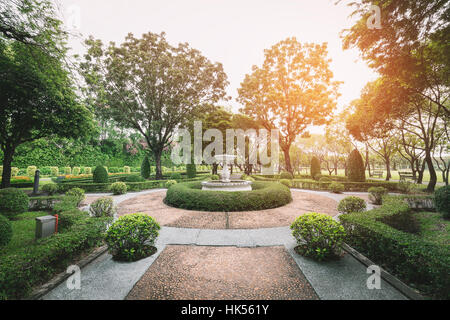 Jardin rond-point décoré par fontaine blanche et arbustes. Avoir l'orange vif la lumière du soleil sur le ciel blanc en matinée. Banque D'Images