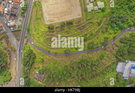 Une vue aérienne de haut en bas montre une route asphaltée sinueuse qui courbe à travers une colline verdoyante et végétalisée. La route se connecte à une route plus droite sur la gauche, près d'un groupe de bâtiments aux toits variés Banque D'Images