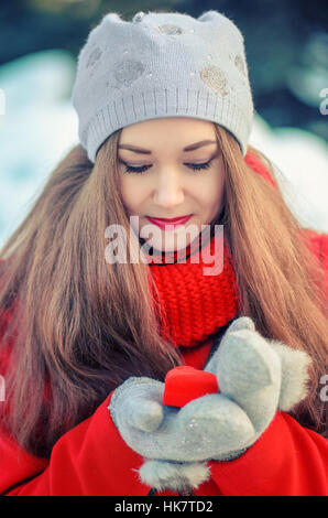 Charmante fille aux cheveux longs dans un manteau rouge et écharpe avec le cadeau dans une boîte en forme de cœur dans les mains. Banque D'Images