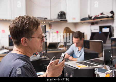 Trois personnes travaillant dans un laboratoire de technologie, avec un homme au premier plan à la recherche à un téléphone cellulaire. Banque D'Images