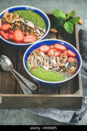 Petit déjeuner sain. Bols smoothie vert avec des fraises, granola, Chia et graines de citrouille, de fruits secs et d'écrou dans le bac en bois sur béton gris retour Banque D'Images