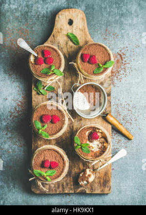 Tiramisu dessert italien fait maison en verres individuels avec des feuilles de menthe, framboises mûres fraîches et de poudre de cacao sur planche de bois rustique gris plus con Banque D'Images