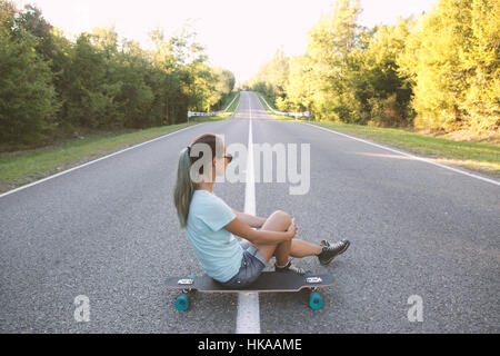 Fille assise sur un longboard et à regarder la route. Banque D'Images