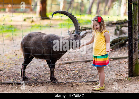 Jolie petite fille en robe colorée à regarder et nourrir les chèvres alpine avec de grandes cornes au zoo par beau jour d'été. Banque D'Images