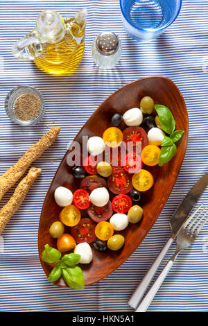 Avec salade de tomates de couleur, de petites mozzarella, olives vertes et les olives noires. Dans un bol ovale en bois. sur nappe à rayures bleu Banque D'Images