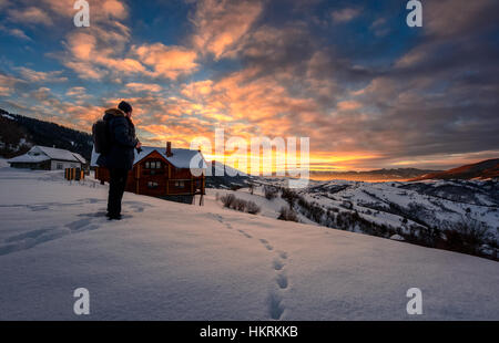 Photographe de paysage se dresse en bordure de colline couverte de neige près du village de maisons dans la région rurale de montagne et regarde sur le lever du soleil d'hiver Banque D'Images