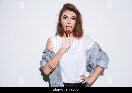 Photo d'une jeune femme drôle vêtue de jeans jacket isolés sur fond blanc tout en mangeant des bonbons. Banque D'Images