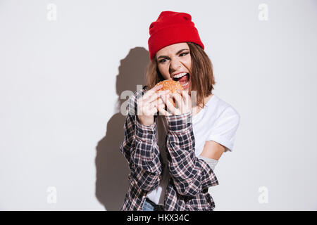 Photo de faim femme vêtue de shirt dans une cage imprimer wearing hat isolés sur fond blanc et holding burger Banque D'Images