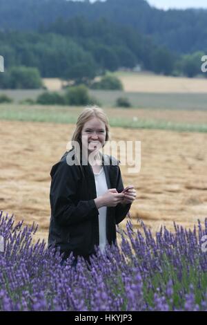 Jeune blonde woman standing in purple champ de lavande avec de l'herbe jaune et vert des arbres en arrière-plan. Banque D'Images