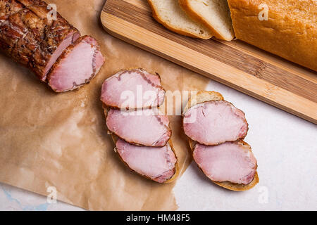 Fumé tranches d'épices et herbes de la viande ou le jambon sur du papier d'emballage brun. Tranches de pain de blé blanc sur la planche à découper en bois. Banque D'Images