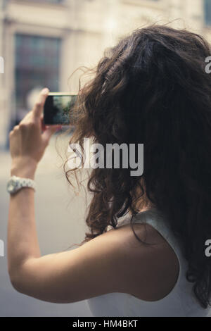 Une jeune fille prendre des photos sur un téléphone intelligent. La jeune fille habillée d'une robe blanche. Elle a les cheveux bruns bouclés. Les rues de photographies de fille Banque D'Images