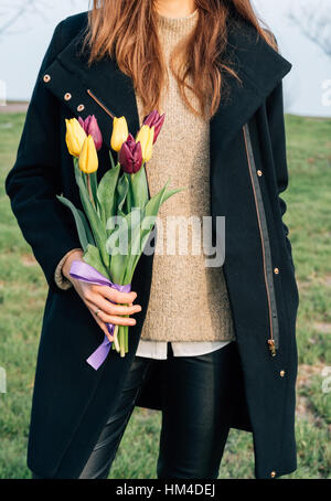 Jeune femme svelte dans un pardessus debout sur un pré vert et tenant un bouquet de tulipes dans la main Banque D'Images