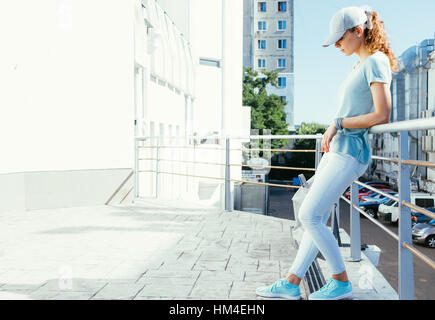 Fille mince d'une casquette et un T-shirt avec un sac à dos dans les mains debout sur un toit plat dans la ville en été Banque D'Images