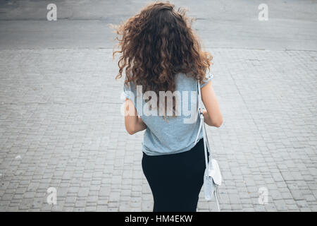 Jeune fille élancée avec de magnifiques cheveux bouclés dans une jupe et sac à main avec une promenade sur un lieu à l'été, la vue de l'arrière Banque D'Images