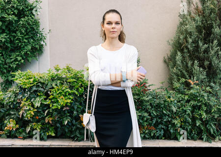 Jeune femme dans une jupe noire et d'un cardigan blanc avec un sac à main tenant un téléphone portable dans sa main à l'extérieur Banque D'Images