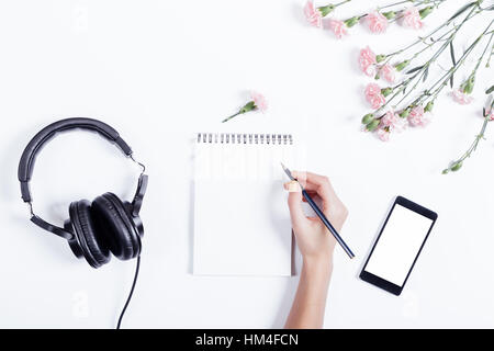 L'écriture main de femme dans un ordinateur portable, téléphone mobile, oreillette et de fleurs sur un fond blanc, vue du dessus Banque D'Images