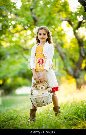 Smiling Girl standing in orchard, holding basket with teddy bear Banque D'Images