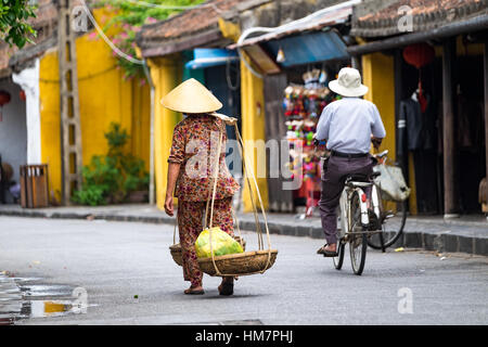 Femme non identifiée en vêtements traditionnels vietnamiens exerçant son buskets avec de la nourriture dans la rue dans la ville de Hoi An. HOI AN, Vietnam Banque D'Images