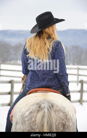 Blonde-haired woman riding a horse Appaloosa gris en hiver, les jeunes cow in casual vêtements occidentaux, un chapeau de cow-boy noir, denim Banque D'Images