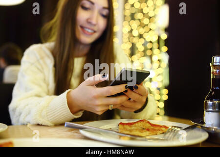 People Ecrire un message sur smart phone dans un café moderne. Portrait de jeune jolie fille assise à une table avec la pizza à l'aide de téléphone mobile. Tonned. Focus sélectif. Banque D'Images